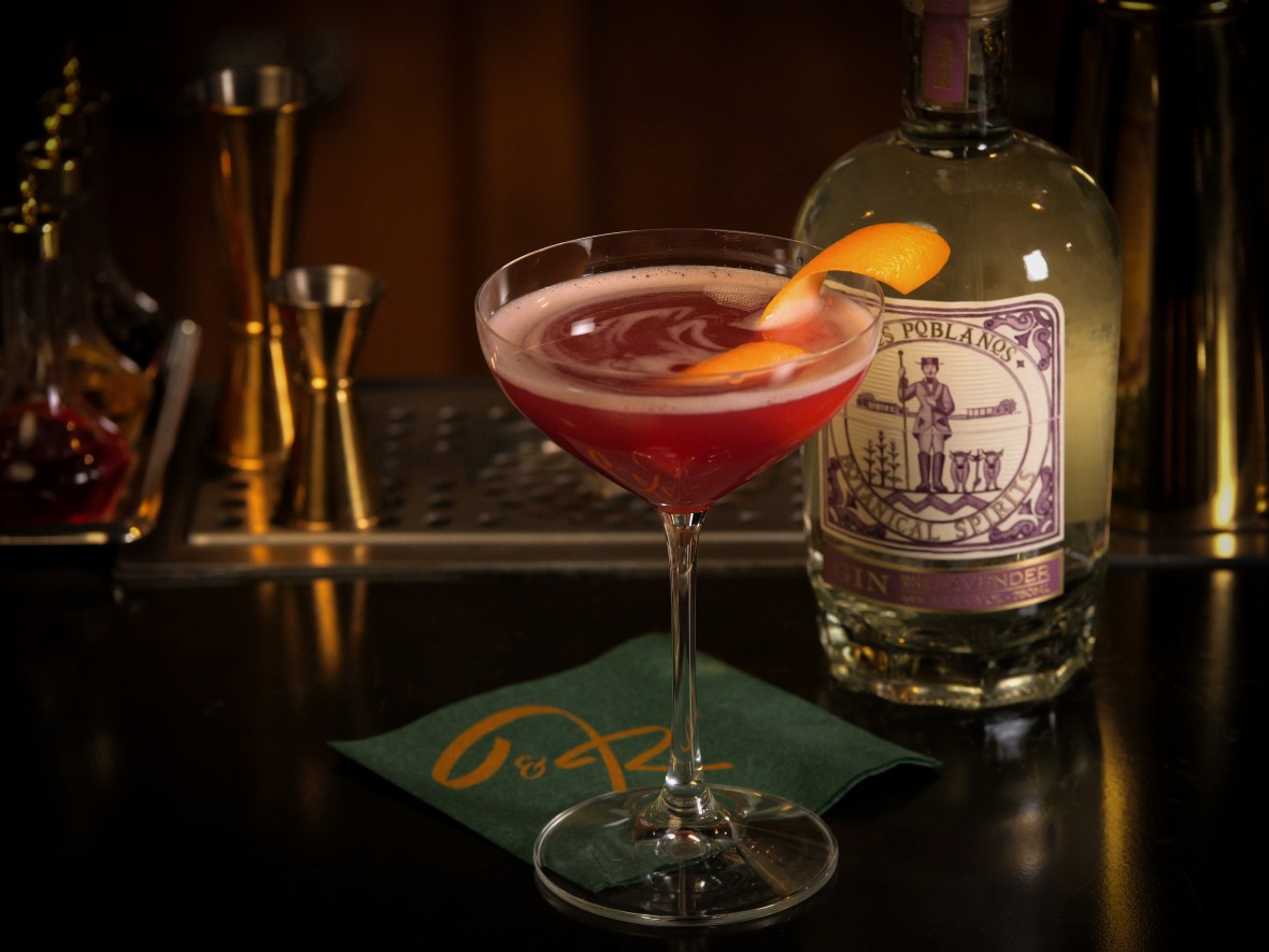A pink cocktail in a martini glass with an orange peel twist sitting on a bar next to a bottle of lavender gin.